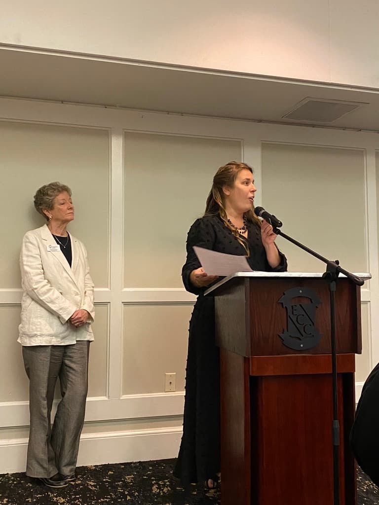 Woman in black dress speaks at a podium while another woman in white listens.