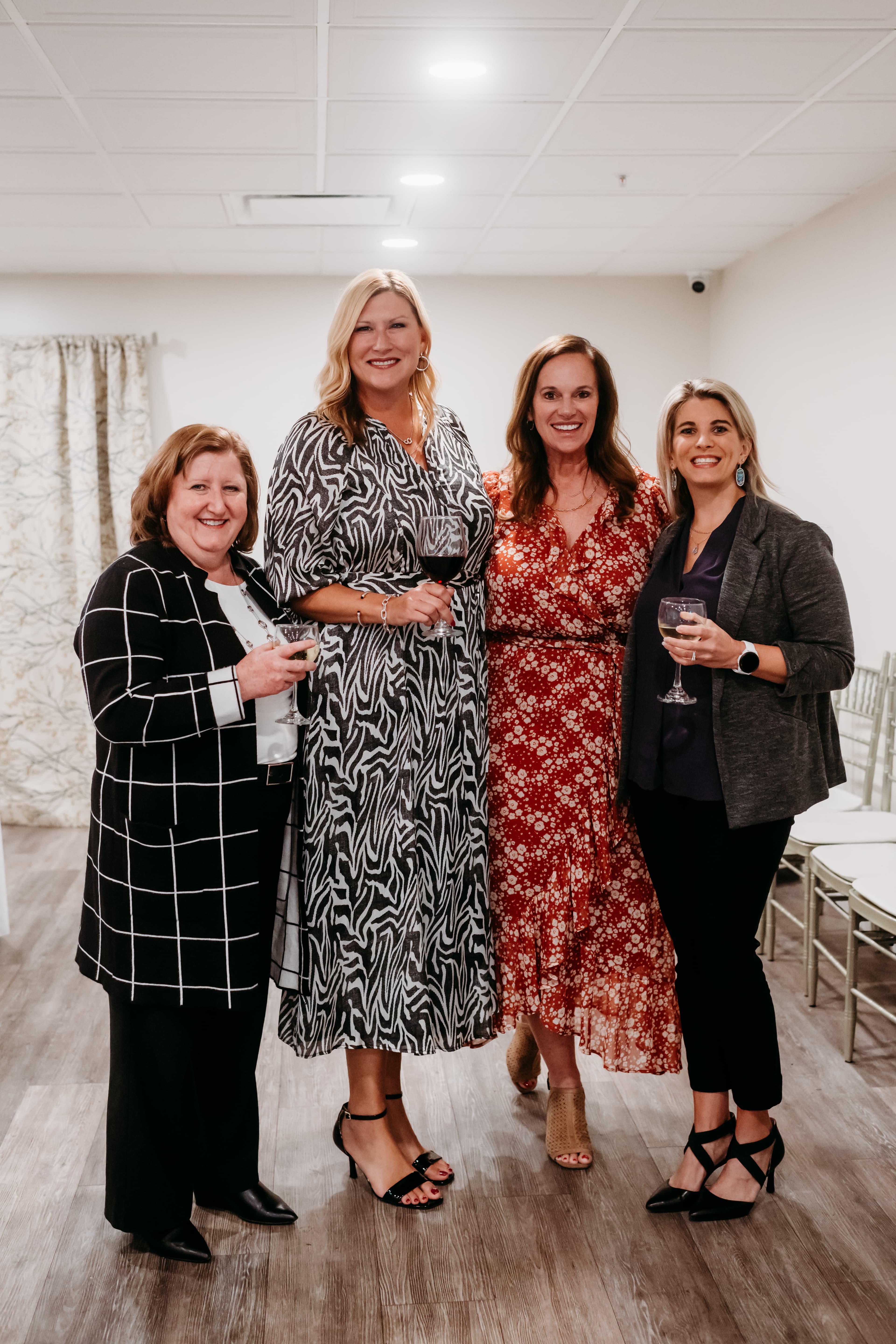 Four smiling women in fashionable outfits hold wine glasses at an indoor social event.