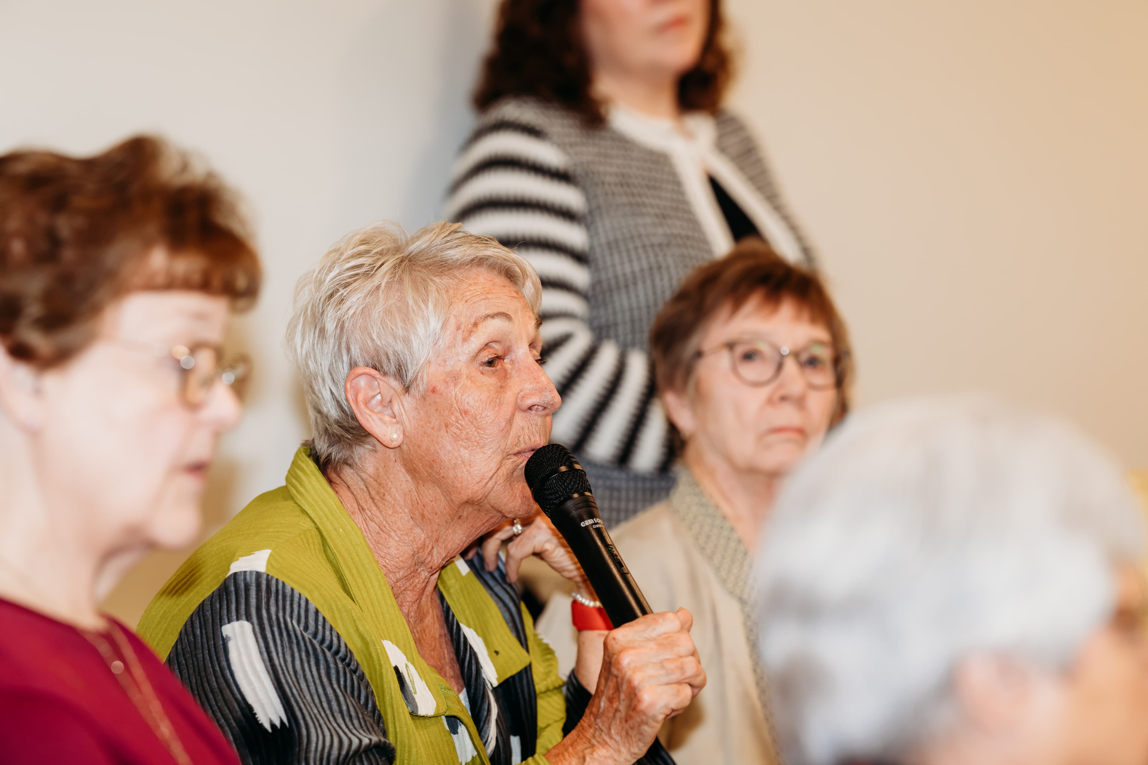 Senior woman with short grey hair speaking into a microphone during a community event.