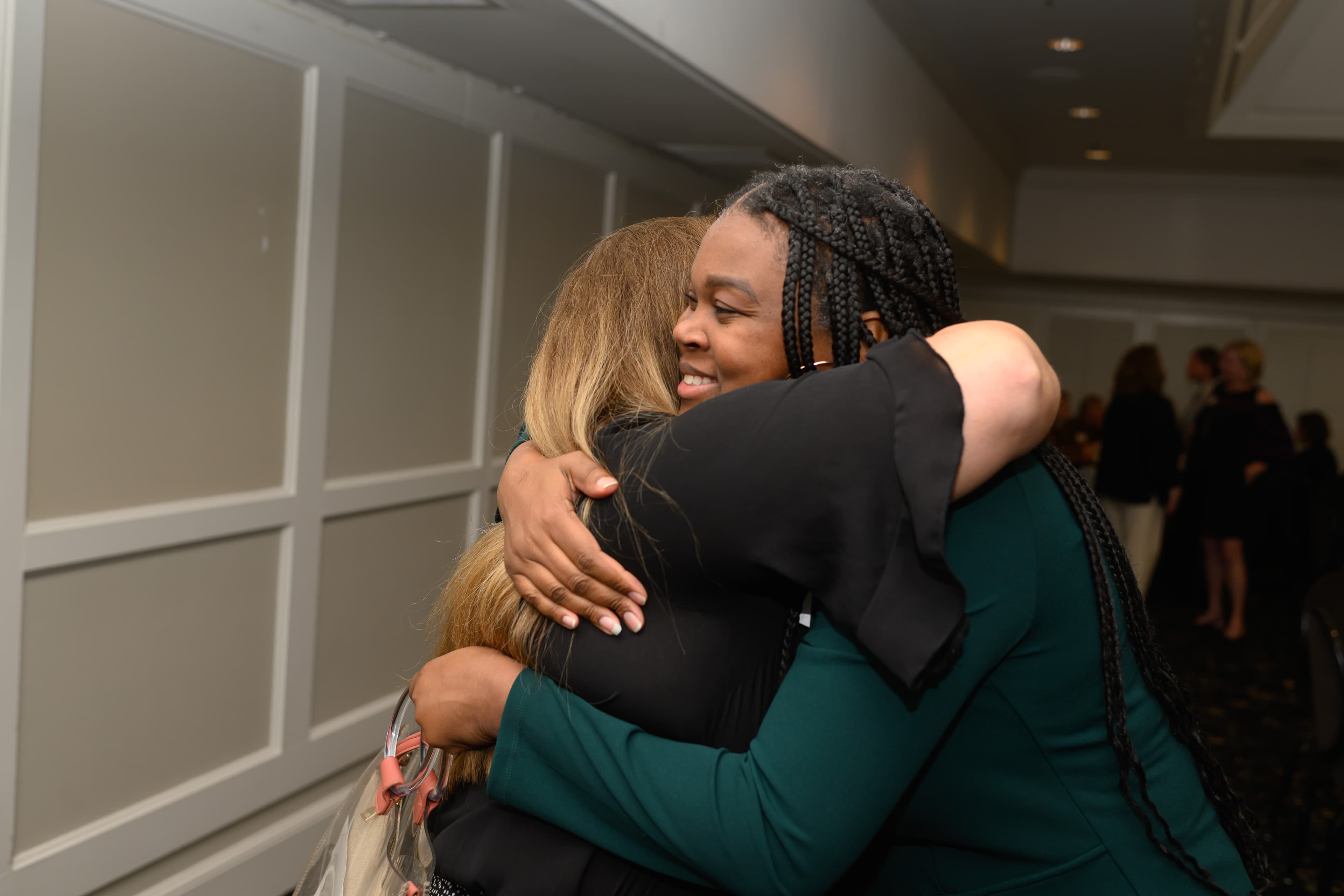 A smiling woman with long braids warmly hugs another woman at a professional event.