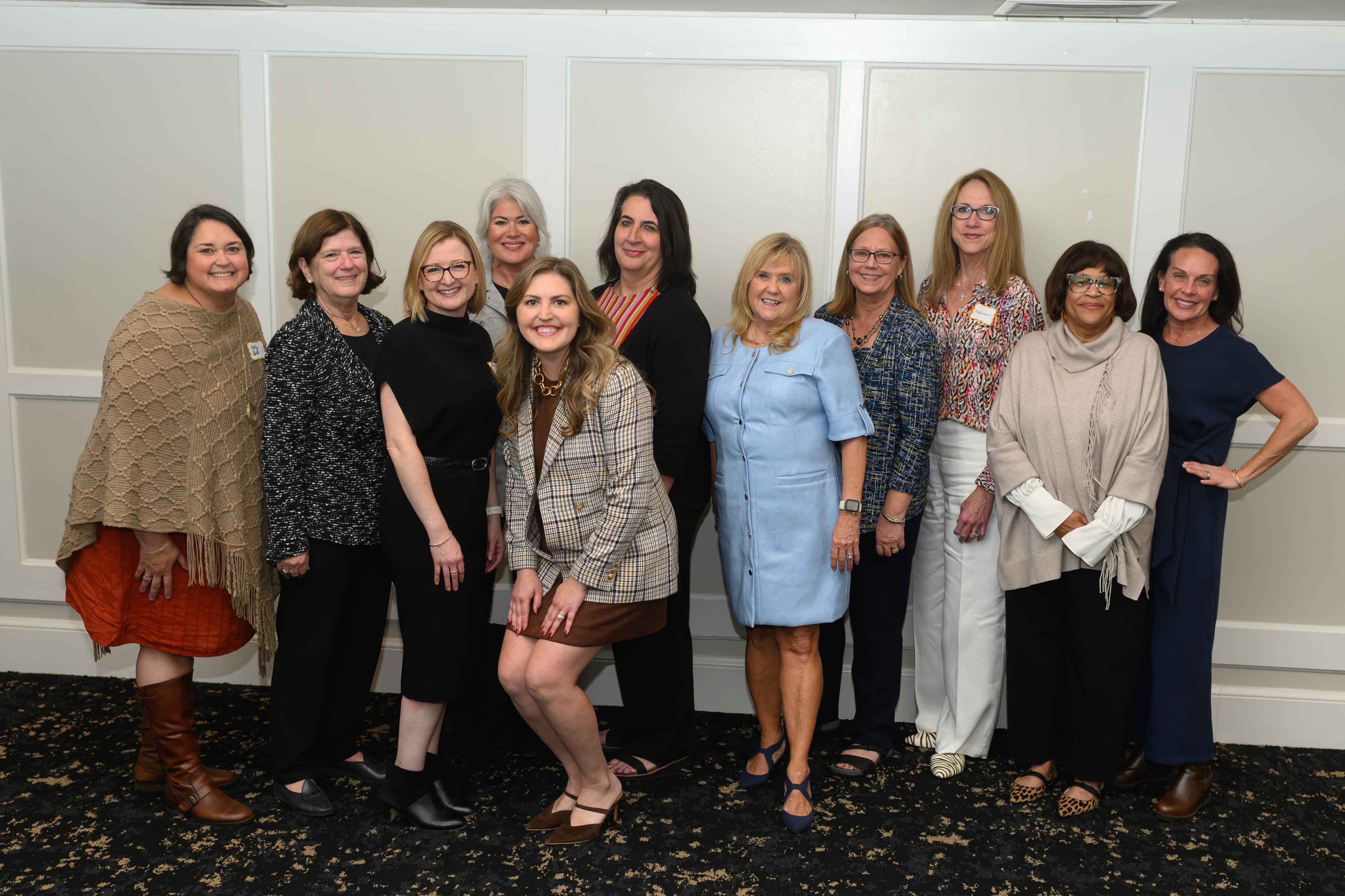 Ten women in professional attire smile and pose together for a group photo indoors.