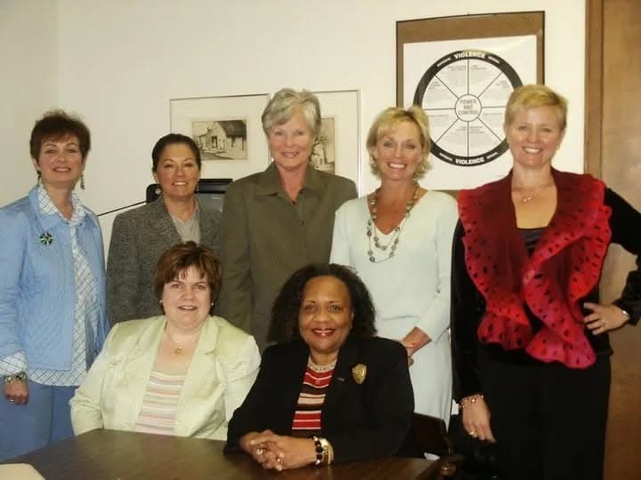 Seven smiling women in professional attire pose for a group portrait in an office.